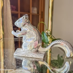 May include: A silver-colored squirrel figurine with a textured tail and a log-shaped base. The squirrel is in a sitting position, with its paws raised. The figurine is displayed on a reflective surface.