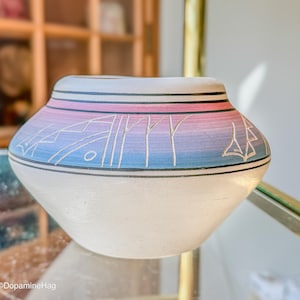 May include: A rounded ceramic pot with a matte white base and a gradient band of pink, purple, and blue across the upper section. The colored area features etched geometric line patterns and is bordered by thin black horizontal lines.