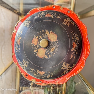 May include: Circular antique serving tray with a black central area and a red, scalloped edge. The tray has floral and gold leaf patterns. A gold-coloured knob is in the centre of the tray.