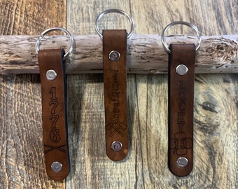 May include: Three brown leather keychains with silver key rings. The keychains are personalized with the text "1776" with crossed axes, "Jones" with crossed axes, and "Lucky 13" with a four-leaf clover.