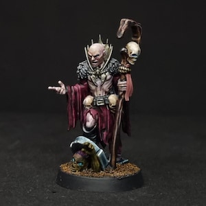 Puede incluir: Una figura en miniatura pintada de un nigromante calvo de piel pálida con una túnica roja oscura, cuello de piel negra y cinturón con calaveras. La figura sostiene un bastón de madera rematado con una calavera sobre una tumba con un esqueleto.