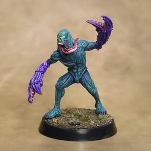 Puede incluir: Una figura en miniatura pintada de una criatura con piel verde azulada, una lengua larga de color rosa y ojos de color verde brillante. La figura tiene grandes manos en forma de garra de color púrpura y se asienta sobre una base negra circular.
