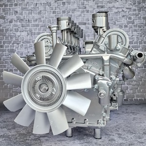 Könnte beinhalten: Ein silberfarbener Motor mit Kolben, einem Lüfter und verschiedenen mechanischen Komponenten. Der Motor wird vor einem grauen Backsteinmauer-Hintergrund präsentiert. Der Lüfter hat mehrere Flügel und ist vorne positioniert.