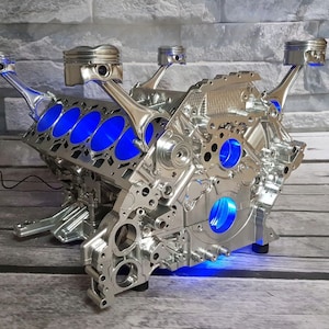 Könnte beinhalten: Ein verchromter Motorblock mit blauen LED-Lichtern, umfunktioniert zu einem einzigartigen Couchtisch. Der Motorblock hat mehrere Zylinder und Pleuel, was ein markantes industrielles Design ergibt.