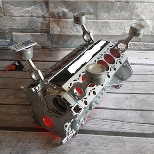 May include: A silver-colored engine block repurposed as a table base, featuring four upright pistons with connecting rods acting as legs. The structure includes red interior lighting and a remote control resting on one of the top piston surfaces.