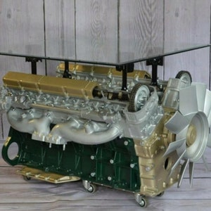 Könnte beinhalten: Ein umgebauter Motorblock mit einer Glasplatte, in Gold und Grün lackiert, dient als Couchtisch. Der Motor hat einen großen Lüfter und Räder an der Unterseite.