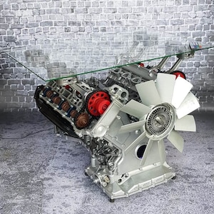 Könnte beinhalten: Ein Couchtisch mit Glasplatte aus einem umgebauten Automotor. Der Motor ist silberfarben lackiert und hat einen großen Ventilator daran befestigt. Der Tisch steht auf einem grauen Betonboden.