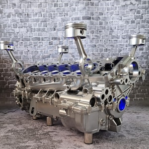 Könnte beinhalten: Ein silberner Motorblock mit vier Kolben und Pleuelstangen, umfunktioniert zu einem Couchtisch. Der Motorblock ist auf einem Metallsockel mit vier Beinen montiert.