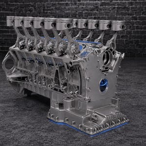 May include: A metallic silver V12 engine block assembly featuring twelve upright pistons connected to their respective rods. The engine structure displays intricate mechanical details, including a cast metal housing, mounting points, and a base pan with a blue accent.