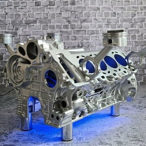 Könnte beinhalten: Ein silberner Motorblock-Couchtisch mit blauen LED-Lichtern. Der Tisch hat vier Beine und zwei Pleuel mit Kolben.