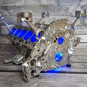 Könnte beinhalten: Ein verchromter Motorblock mit blauen LED-Lichtern, umfunktioniert zu einem einzigartigen Couchtisch. Der Motorblock hat mehrere Zylinder und Pleuel, was ein markantes industrielles Design ergibt.