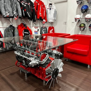 Könnte beinhalten: Ein Couchtisch mit Glasplatte aus einem wiederverwendeten Motorblock. Der Tisch hat ein rotes Untergestell und eine klare Glasplatte. Der Tisch steht in einem Raum mit roten Stühlen und Motorradbekleidung an den Wänden.