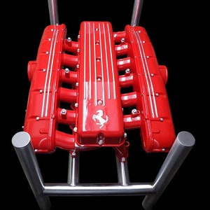 Könnte beinhalten: Ein roter Ferrari-Motorblock, der auf einem Edelstahlständer montiert ist. Der Motorblock hat ein silbernes Ferrari-Logo auf der Oberseite.