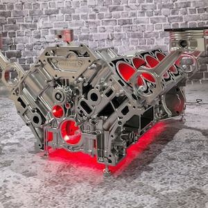 Könnte beinhalten: Ein polierter silberner Motorblock mit roter LED-Beleuchtung. Der Motorblock hat Kolben und Pleuelstangen. Der Hintergrund ist eine graue Backsteinmauer.