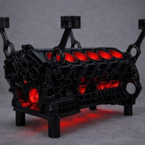 Könnte beinhalten: Ein schwarzer Tisch im Industriestil, gefertigt aus einem Motorblock. Der Tisch verfügt über eine rote Innenbeleuchtung und nach oben ragende Kolben. Er wird von vier zylindrischen Beinen getragen und ist ein einzigartiges Möbelstück. Maße in Zentimetern.