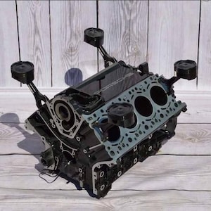 Könnte beinhalten: Ein schwarzer Porsche-Motorblock mit ausgefahrenen Kolben. Der Motorblock hat eine silberfarbene Dichtung, und das Wort "PORSCHE" ist oben sichtbar. Der Hintergrund ist eine helle Holzmaserung.