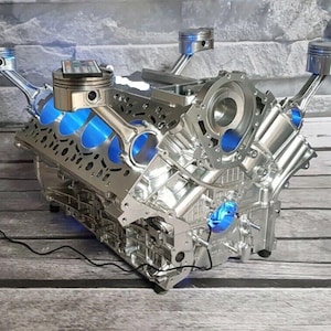 May include: A silver-colored V-engine block repurposed as a table base, featuring four metallic pistons extending upward. The engine block has a polished finish with bright blue interior lighting and a black power cord extending from the side.