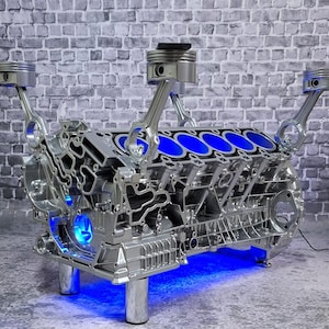 Könnte beinhalten: Ein silberner Motorblock mit blauer LED-Beleuchtung. Der Motorblock hat vier Kolben und Pleuelstangen. Der Motorblock ist auf vier silbernen Beinen montiert. Der Hintergrund ist eine graue Backsteinmauer.