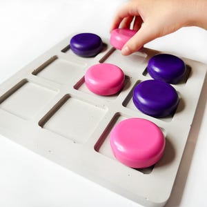 May include: A gray tic-tac-toe board with pink and purple game pieces. A hand is placing a pink piece on the board. The game pieces are round and fit into the square spaces on the board.