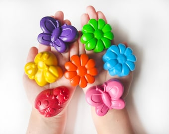 May include: A collection of colorful, shaped crayons in the form of a butterfly, flower, ladybug, and bee. The crayons are in shades of purple, green, blue, orange, yellow, pink, and red. The crayons are held in open hands.