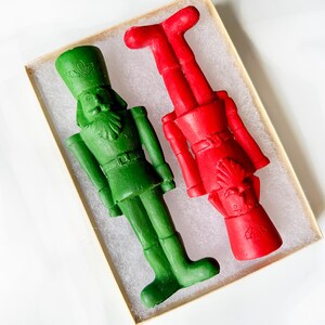 May include: Two decorative nutcracker figurines, one green and one red, are displayed in a rectangular box. The green nutcracker is upright, while the red one is inverted. Both have detailed uniforms and hats, perfect for holiday decor.