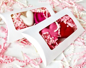 May include: Two white pillow boxes with heart-shaped items inside. One box contains a white heart and a pink heart, the other contains a pink and a red heart. The boxes are surrounded by pink and white paper shreds.