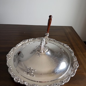 May include: A silver-plated serving dish with a domed lid and ornate, scalloped edges. The lid features a decorative handle with a dark brown wooden grip. The dish is likely used for serving food and has a classic, elegant design.
