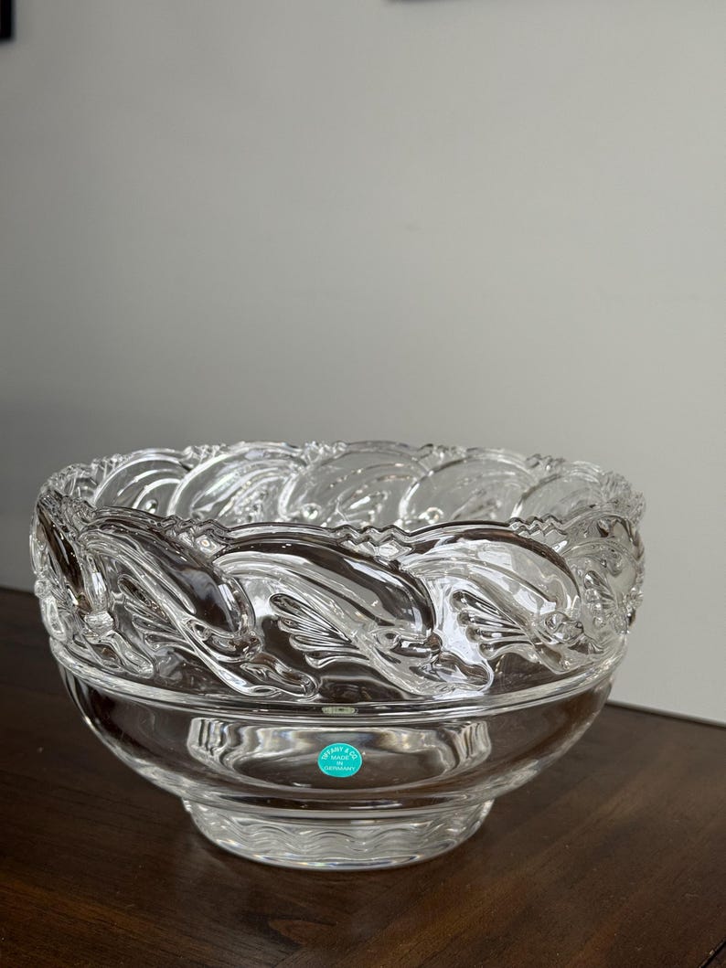 May include: A clear glass bowl with a decorative dolphin design around the rim. The bowl has a round base and a wide opening. A small, round, blue and white label is affixed to the bowl.