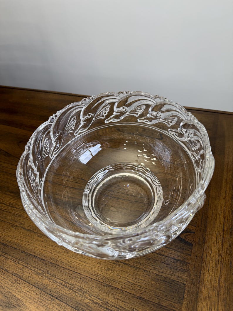 May include: Clear glass serving bowl with a decorative dolphin design around the rim. The bowl has a smaller, circular bowl in the center. The bowl is sitting on a wooden surface.