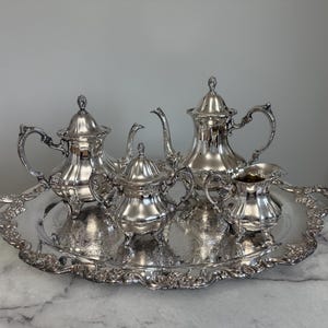 May include: A silver tea set on a decorative tray. The set includes a teapot, creamer, sugar bowl, and two other vessels. The tray and tea set have ornate detailing and are displayed on a marble-topped table.