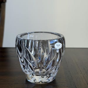 May include: A clear crystal glass with a textured, starburst-like pattern. The glass has a rounded shape and a wide opening. A small white label with text is affixed to the side. The glass sits on a dark wooden surface.
