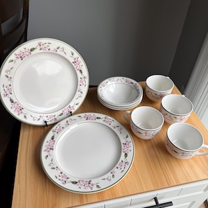 May include: A collection of white ceramic dinnerware with a floral pattern. The set includes plates, bowls, and teacups. The floral design features pink blossoms and green accents. The pieces are arranged on a wooden surface.