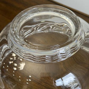 May include: A clear glass bowl with a wide, wavy rim. The bowl has a smooth, reflective surface and appears to be empty. The text "TIFFANY & CO" is visible on the rim. The bowl sits on a wooden surface.