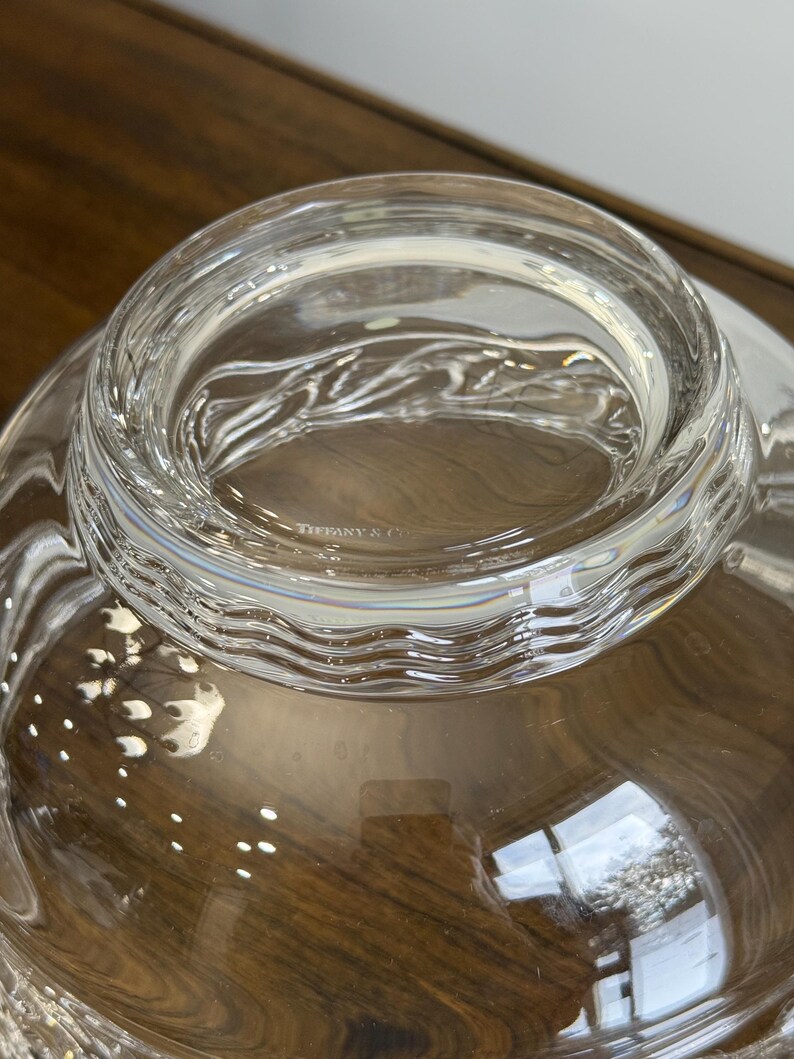 May include: A clear glass bowl with a wide, wavy rim. The bowl has a smooth, reflective surface and appears to be empty. The text "TIFFANY & CO" is visible on the rim. The bowl sits on a wooden surface.
