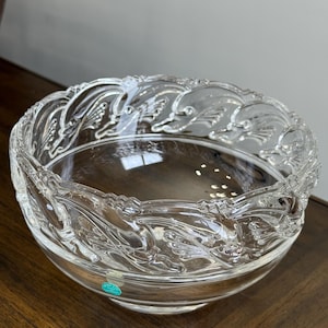 May include: A clear glass bowl with a decorative, raised leaf pattern around the rim. The bowl is round and sits on a dark wooden surface. The bowl is likely for decorative or serving purposes.