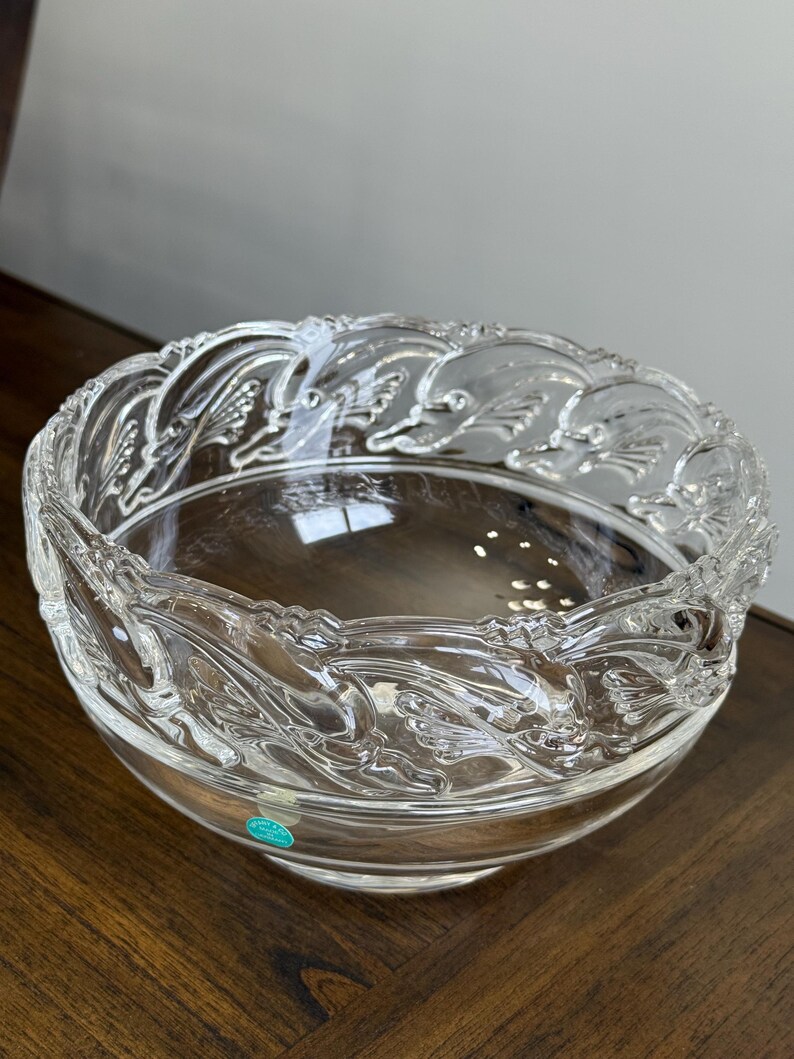 May include: A clear glass bowl with a decorative, raised leaf pattern around the rim. The bowl is round and sits on a dark wooden surface. The bowl is likely for decorative or serving purposes.