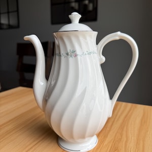 May include: White porcelain coffee pot with a lid and a curved handle. The pot features a spiral design and is decorated with a floral pattern in pink and green. A silver trim accents the base and the lid.