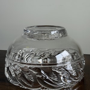 May include: A clear glass bowl with a lid. The bowl features a detailed relief design of stylized waves and mythical creatures around the base. The lid has a wavy pattern. The bowl sits on a dark wooden surface.