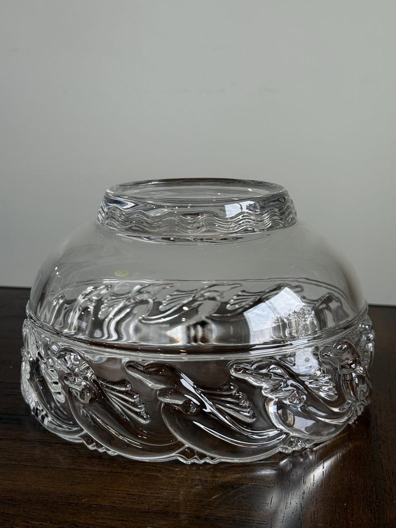 May include: A clear glass bowl with a lid. The bowl features a detailed relief design of stylized waves and mythical creatures around the base. The lid has a wavy pattern. The bowl sits on a dark wooden surface.