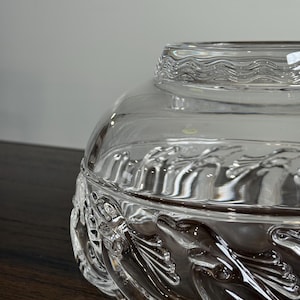 May include: A clear glass decorative bowl with a lid. The bowl features a detailed leaf and vine pattern around the base. The lid has a wavy, textured design. The bowl sits on a dark wooden surface.