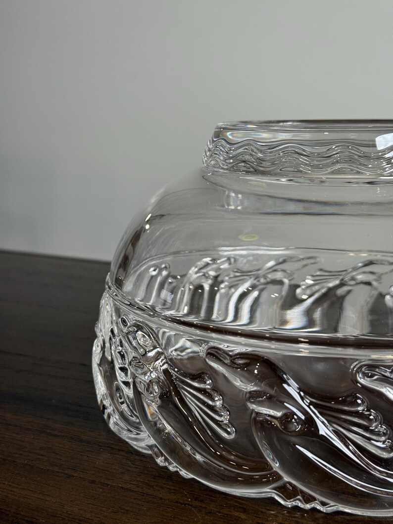 May include: A clear glass decorative bowl with a lid. The bowl features a detailed leaf and vine pattern around the base. The lid has a wavy, textured design. The bowl sits on a dark wooden surface.