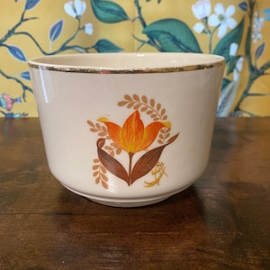 Könnte beinhalten: Ein weißer Keramiktopf mit goldfarbenem Rand und einem Blumendesign mit einer orangefarbenen Tulpe mit braunen Blättern.