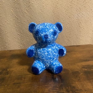 May include: A blue and white ceramic teddy bear figurine. The bear is sitting with its legs crossed and has a speckled blue and white glaze.