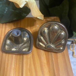 May include: Two antique brass drawer pulls. One pull features a shell design, while the other has a threaded post for attachment. The pulls are a warm gold color and are set against a light wood surface.