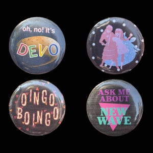 May include: Four circular pinback buttons featuring music-themed graphics. One reads 'oh, no! it's DEVO', another shows two figures in a polka dot pattern, one displays 'OINGO BOINGO' in stylized text, and the last says 'ASK ME ABOUT NEW WAVE'.