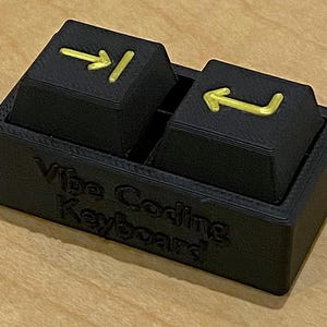 May include: A black keyboard with two keys, each with a gold arrow symbol. The keyboard base has the text "Vibe Coding Keyboard" embossed on the front. The keys are rectangular and sit in a rectangular base.