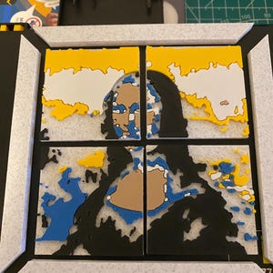 May include: A 3D-printed artwork depicting a fragmented portrait, featuring yellow, white, blue, and brown elements against a black background. The artwork is divided into four sections, framed in white.