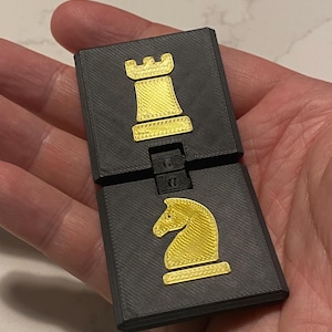 May include: A black, hinged chess piece with a gold rook and knight design. The chess piece is made of a textured material and is held open, revealing the two chess pieces. The rook and knight are in a gold color.
