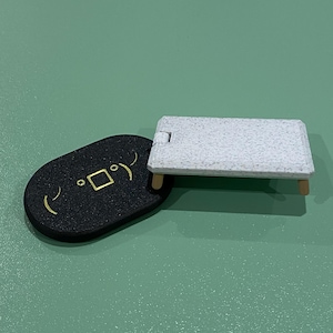 May include: A black, oval-shaped object with a gold square and curved lines, next to a small, rectangular white table with wooden legs. The objects are on a green surface.