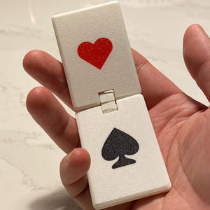 Puede incluir: Un objeto blanco con forma de carta de juego con bisagras. Un lado presenta un corazón rojo, el otro un símbolo de pica negro. El objeto se mantiene abierto, revelando ambas caras de la carta. El artículo parece estar hecho de un material texturizado, posiblemente plástico.